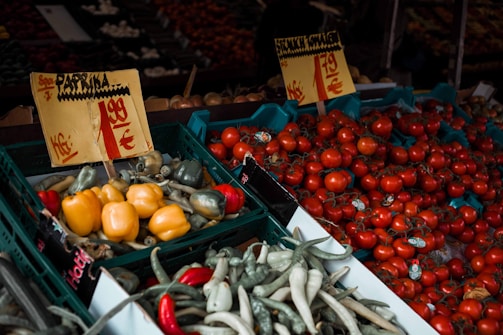 A vibrant display of fresh vegetables including tomatoes, bell peppers, and chili peppers, arranged in crates at a market stall. The vegetables are accompanied by handwritten price signs indicating their cost per kilogram.