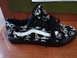 A black sneaker featuring cartoon character designs in white. The shoe has black laces and a distinctive white stripe. The design includes speech bubbles with phrases like 'Good Grief.' The sneaker rests on a wooden surface, with a green and brown background.