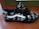 A black sneaker featuring cartoon character designs in white. The shoe has black laces and a distinctive white stripe. The design includes speech bubbles with phrases like 'Good Grief.' The sneaker rests on a wooden surface, with a green and brown background.