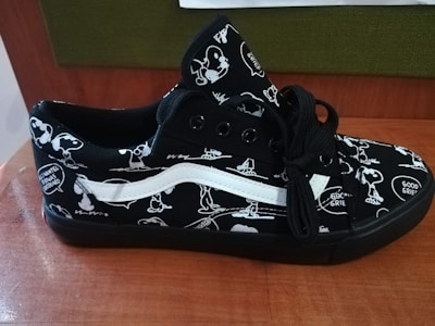 A black sneaker featuring cartoon character designs in white. The shoe has black laces and a distinctive white stripe. The design includes speech bubbles with phrases like 'Good Grief.' The sneaker rests on a wooden surface, with a green and brown background.