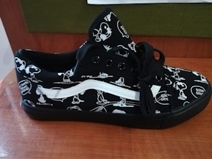 A black sneaker featuring cartoon character designs in white. The shoe has black laces and a distinctive white stripe. The design includes speech bubbles with phrases like 'Good Grief.' The sneaker rests on a wooden surface, with a green and brown background.