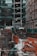 A busy urban construction site with open trenches surrounded by orange barriers and green netting. Large pipes and cables are visible beneath the ground, with sandbags and piles of rubble scattered around. Adjacent high-rise buildings with glass facades and scaffolding are seen in the background. Streets are lined with parked vehicles, and a few urban elements like a tree and signage are present.