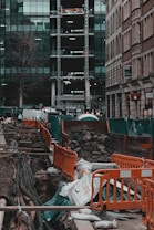 A busy urban construction site with open trenches surrounded by orange barriers and green netting. Large pipes and cables are visible beneath the ground, with sandbags and piles of rubble scattered around. Adjacent high-rise buildings with glass facades and scaffolding are seen in the background. Streets are lined with parked vehicles, and a few urban elements like a tree and signage are present.