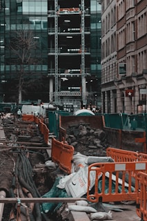 A busy urban construction site with open trenches surrounded by orange barriers and green netting. Large pipes and cables are visible beneath the ground, with sandbags and piles of rubble scattered around. Adjacent high-rise buildings with glass facades and scaffolding are seen in the background. Streets are lined with parked vehicles, and a few urban elements like a tree and signage are present.
