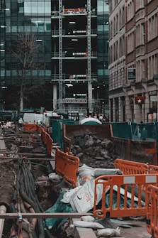A busy urban construction site with open trenches surrounded by orange barriers and green netting. Large pipes and cables are visible beneath the ground, with sandbags and piles of rubble scattered around. Adjacent high-rise buildings with glass facades and scaffolding are seen in the background. Streets are lined with parked vehicles, and a few urban elements like a tree and signage are present.