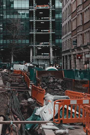A busy urban construction site with open trenches surrounded by orange barriers and green netting. Large pipes and cables are visible beneath the ground, with sandbags and piles of rubble scattered around. Adjacent high-rise buildings with glass facades and scaffolding are seen in the background. Streets are lined with parked vehicles, and a few urban elements like a tree and signage are present.