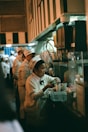 A woman in a white uniform with a hat works behind a counter, preparing drinks or food. There are several other people wearing similar uniforms lined up, indicating a busy environment like a kitchen or a food stand.