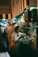 A woman in a white uniform with a hat works behind a counter, preparing drinks or food. There are several other people wearing similar uniforms lined up, indicating a busy environment like a kitchen or a food stand.