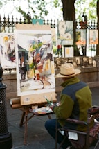 An artist is seated on a folding chair, working on a colorful painting placed on an easel in an outdoor setting. The painting depicts an abstract urban scene with buildings and trees. The artist is wearing a straw hat and a green jacket, and a palette with paint tubes is on a table beside them. Various artworks are displayed on a fence in the background, suggesting a street art fair or exhibition.