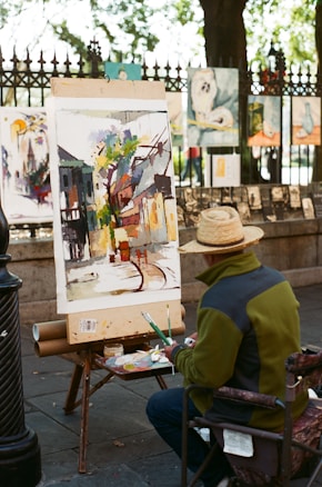 An artist is seated on a folding chair, working on a colorful painting placed on an easel in an outdoor setting. The painting depicts an abstract urban scene with buildings and trees. The artist is wearing a straw hat and a green jacket, and a palette with paint tubes is on a table beside them. Various artworks are displayed on a fence in the background, suggesting a street art fair or exhibition.