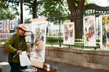 An artist is outdoors, painting on a canvas set up on an easel. He is wearing a straw hat and a green jacket. Various colorful paintings are displayed on a fence behind him, with a park visible in the background. The scene is illuminated by natural daylight, creating a vibrant and lively atmosphere.