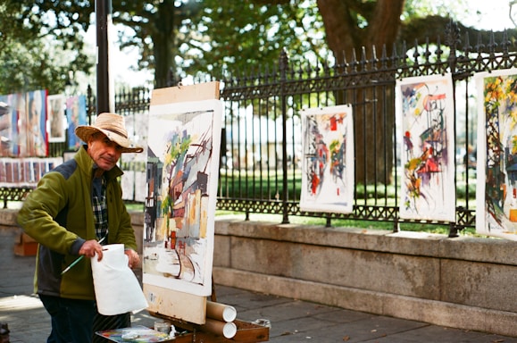 An artist is outdoors, painting on a canvas set up on an easel. He is wearing a straw hat and a green jacket. Various colorful paintings are displayed on a fence behind him, with a park visible in the background. The scene is illuminated by natural daylight, creating a vibrant and lively atmosphere.
