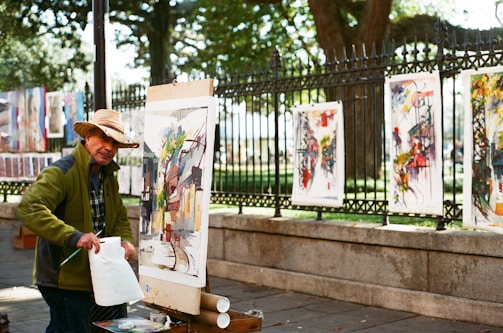 An artist is outdoors, painting on a canvas set up on an easel. He is wearing a straw hat and a green jacket. Various colorful paintings are displayed on a fence behind him, with a park visible in the background. The scene is illuminated by natural daylight, creating a vibrant and lively atmosphere.