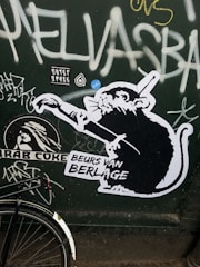 A graffiti-covered wall features a black and white stencil image of a rat holding a paintbrush, with text reading 'BEURS VAN BERLAGE' and various tags and stickers surrounding it. The dark green background is adorned with additional graffiti in white and yellow, and part of a bicycle wheel is visible at the bottom left corner.