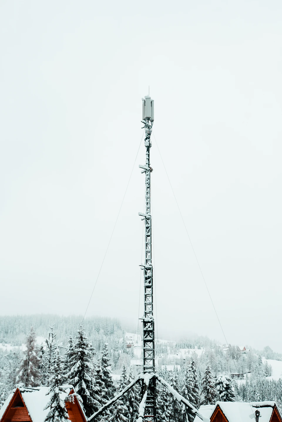 The image depicts a snow-covered mountain with a tall, gray metal tower reaching towards the sky on its left side. The tower is equipped with antennas and wires that extend upwards to connect to various communication systems. In the background, there are several houses nestled among the trees, their rooftops blanketed in snow.

The scene conveys a sense of tranquility and isolation, as if time has come to a halt on this snowy mountain peak. The image is taken from a low angle, emphasizing the height of the tower and the vastness of the surrounding landscape.
