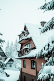 A snow-covered chalet-style house with a sloped roof and wooden balconies is surrounded by pine trees heavily laden with snow. The background features more snow-covered houses and tall pine trees. The overall scene is serene and wintry.