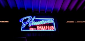A brightly illuminated neon sign with the word 'Hometown' in blue cursive script dominates the image. Below it, 'BARBECUE' is displayed in red block letters, followed by smaller text offering 'BEER', 'COCKTAILS', and 'WINE'. The background is dark, enhancing the vivid glow of the neon lights. The sign is set against a grid-like pattern and surrounded by a ceiling of exposed beams.