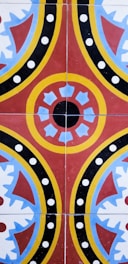 A colorful geometric pattern consisting of circular and semi-circular shapes with red, yellow, blue, and black colors arranged symmetrically. The design features a central circular motif with surrounding crescent and spiked shapes, accented by polka dots.