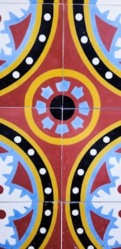 A colorful geometric pattern consisting of circular and semi-circular shapes with red, yellow, blue, and black colors arranged symmetrically. The design features a central circular motif with surrounding crescent and spiked shapes, accented by polka dots.