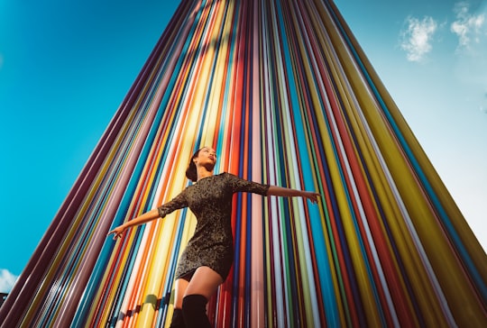 woman wearing black short-sleeved mini dress raising both hands while standing near multicolored high-rise building under blue and white sky in La Défense France