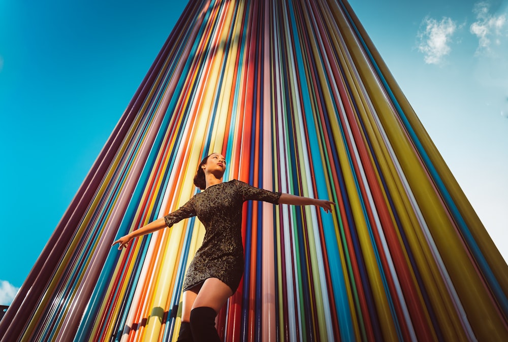 woman wearing black short-sleeved mini dress raising both hands while standing near multicolored high-rise building under blue and white sky