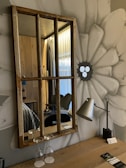 A rustic wooden-framed mirror hangs on a wall painted with a large, abstract flower. Below the mirror, two upside-down wine glasses rest on a wooden table alongside a modern metal desk lamp. The room reflected in the mirror features wooden paneling, a hint of a bed with striped bedding, and closed curtains of a translucent fabric.