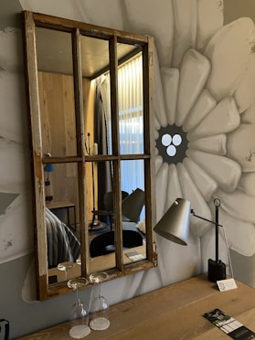 A rustic wooden-framed mirror hangs on a wall painted with a large, abstract flower. Below the mirror, two upside-down wine glasses rest on a wooden table alongside a modern metal desk lamp. The room reflected in the mirror features wooden paneling, a hint of a bed with striped bedding, and closed curtains of a translucent fabric.