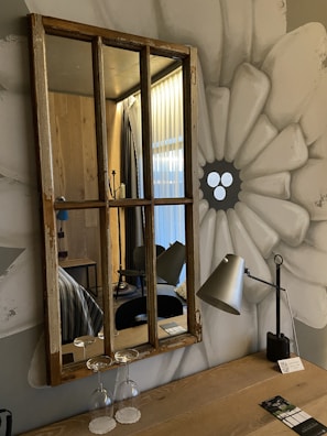 A rustic wooden-framed mirror hangs on a wall painted with a large, abstract flower. Below the mirror, two upside-down wine glasses rest on a wooden table alongside a modern metal desk lamp. The room reflected in the mirror features wooden paneling, a hint of a bed with striped bedding, and closed curtains of a translucent fabric.