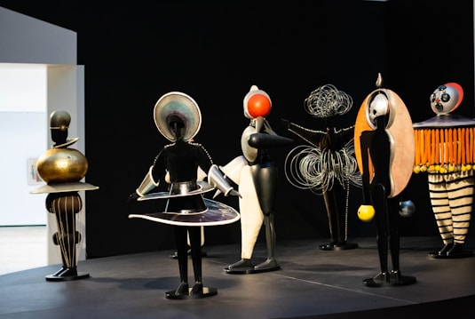 A group of abstract sculptures stand on a platform, resembling futuristic human-like figures. Each sculpture is uniquely designed with intricate patterns, metal elements, and geometric shapes. The background is dark, accentuating the vibrant colors of the figures.