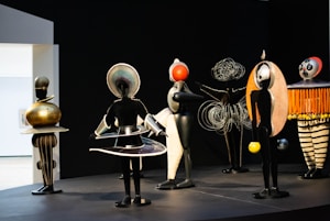 A group of abstract sculptures stand on a platform, resembling futuristic human-like figures. Each sculpture is uniquely designed with intricate patterns, metal elements, and geometric shapes. The background is dark, accentuating the vibrant colors of the figures.
