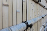 A close-up view of a wall made from vertically aligned bamboo sticks, tied together with black rope. The bamboo appears weathered and has a natural beige color with some grayish tones. The texture of the bamboo and the rope is clearly visible, creating a rustic and natural aesthetic.