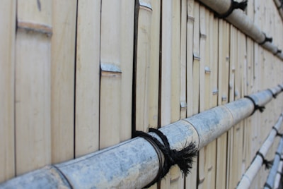 A close-up view of a wall made from vertically aligned bamboo sticks, tied together with black rope. The bamboo appears weathered and has a natural beige color with some grayish tones. The texture of the bamboo and the rope is clearly visible, creating a rustic and natural aesthetic.