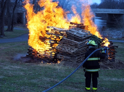 A firefighter dressed in protective gear is attempting to extinguish a large blaze consuming a stack of wooden pallets outdoors. The fire emits intense orange and yellow flames, contrasting with the surrounding grassy area and leafless trees. A body of water is visible in the distance, along with a bridge.