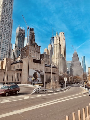 A cityscape featuring various tall skyscrapers under a clear blue sky. Construction cranes are visible among the buildings, indicating ongoing development. The street below has vehicles, and an advertisement billboard is prominently displayed on one of the buildings.