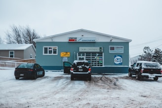 A small convenience store named Chubb's Convenience is situated in a snowy landscape. Several cars are parked in front of the store on a snow-covered lot. Signs on the storefront indicate the sale of lottery tickets and the presence of an ATM. The building is painted in shades of blue with signage for Pepsi and Liquor Express.