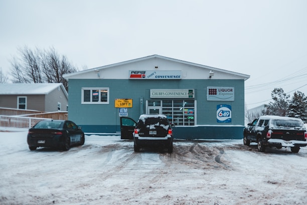 A small convenience store named Chubb's Convenience is situated in a snowy landscape. Several cars are parked in front of the store on a snow-covered lot. Signs on the storefront indicate the sale of lottery tickets and the presence of an ATM. The building is painted in shades of blue with signage for Pepsi and Liquor Express.