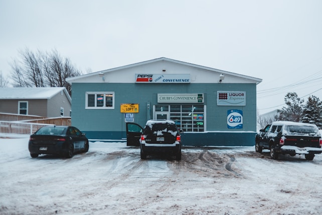 A small convenience store named Chubb's Convenience is situated in a snowy landscape. Several cars are parked in front of the store on a snow-covered lot. Signs on the storefront indicate the sale of lottery tickets and the presence of an ATM. The building is painted in shades of blue with signage for Pepsi and Liquor Express.
