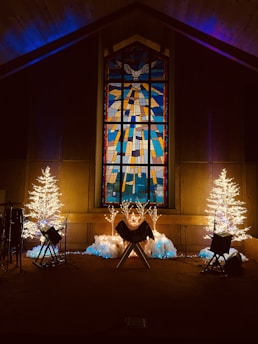 In a dimly lit room, a large stained glass window featuring a dove at the top center is flanked by two illuminated artificial trees. Below the window, a manger-like structure is surrounded by faux snow and blue lights. Musical instruments stand nearby, suggesting a festive or religious setting.