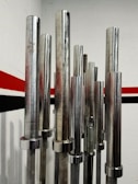 Several metallic barbell bars are standing vertically in a room with a red and black stripe running along the white wall.