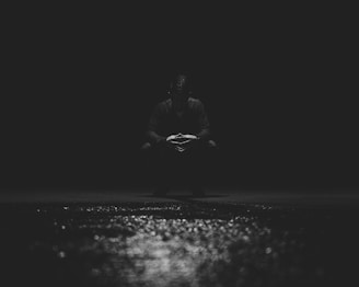 A figure is seated in a dark, shadowy setting with a reflective surface in the foreground. The person appears to be in deep thought, adding a mysterious and introspective mood to the scene.