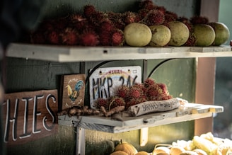 A rustic fruit stand with shelves displaying tropical fruits including rambutan and green papayas. A wooden sign reads 'Bananas for sale' next to a Hawaii license plate. Warm sunlight creates a cozy atmosphere while a colorful rooster illustration adds charm.