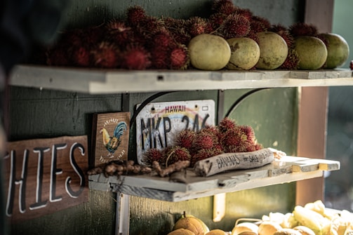 A rustic fruit stand with shelves displaying tropical fruits including rambutan and green papayas. A wooden sign reads 'Bananas for sale' next to a Hawaii license plate. Warm sunlight creates a cozy atmosphere while a colorful rooster illustration adds charm.