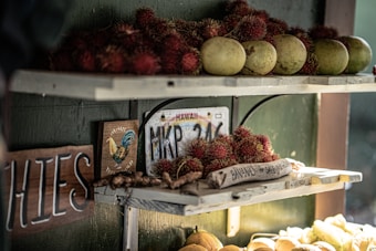 A rustic fruit stand with shelves displaying tropical fruits including rambutan and green papayas. A wooden sign reads 'Bananas for sale' next to a Hawaii license plate. Warm sunlight creates a cozy atmosphere while a colorful rooster illustration adds charm.