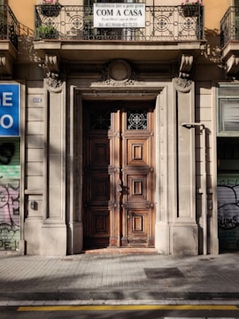 A large, ornate wooden door is set into an intricately carved stone facade. Above the door is a balcony with an iron railing and potted flowers. The building appears to be an old, possibly historical structure with classical architectural elements. A sign in a foreign language is visible, indicating it may be a business or service.