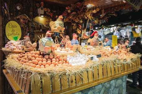 A festive market stall displays a variety of fresh eggs piled on straw, surrounded by decorative ceramic chickens. The wooden stall is adorned with rustic decorations, creating a quaint village market atmosphere.
