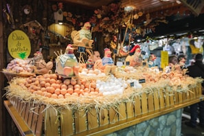 A festive market stall displays a variety of fresh eggs piled on straw, surrounded by decorative ceramic chickens. The wooden stall is adorned with rustic decorations, creating a quaint village market atmosphere.