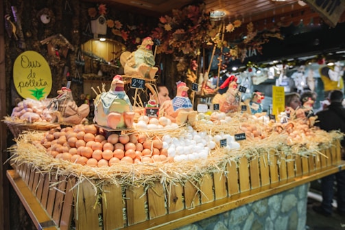 A festive market stall displays a variety of fresh eggs piled on straw, surrounded by decorative ceramic chickens. The wooden stall is adorned with rustic decorations, creating a quaint village market atmosphere.