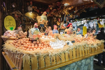 A festive market stall displays a variety of fresh eggs piled on straw, surrounded by decorative ceramic chickens. The wooden stall is adorned with rustic decorations, creating a quaint village market atmosphere.