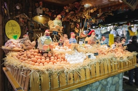 A festive market stall displays a variety of fresh eggs piled on straw, surrounded by decorative ceramic chickens. The wooden stall is adorned with rustic decorations, creating a quaint village market atmosphere.