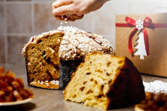 A sliced panettone on a table, with a hand sprinkling white sugar granules on top. The panettone has a golden-brown crust with visible chunks of dried fruit. In the background, there is a brown gift box with a red ribbon, featuring a decorative white snowflake and tree design.